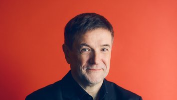 Autor vor rotem Huntergrund, lächelnd, schwarzes Hemd, kurze braune Haare