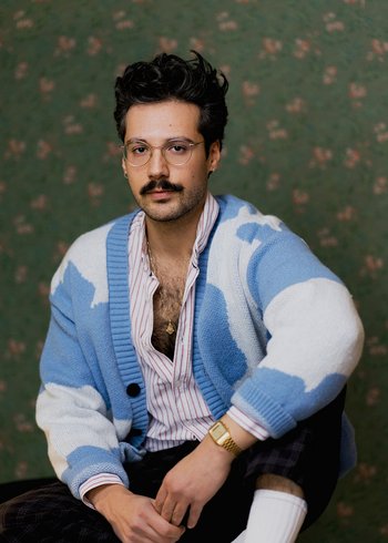 junger Mann in blau-weißem Cardigan, dunkle Haare und dunkler SChnurrbart, silberne Brille