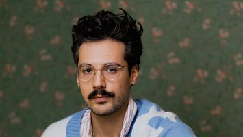 junger Mann in blau-weißem Cardigan, dunkle Haare und dunkler SChnurrbart, silberne Brille