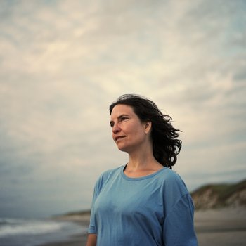 Autorin im Halbprofil, blaues Kurzarmshirt, wehende schulterlange, braune Haare, vor bedecktem Himmel