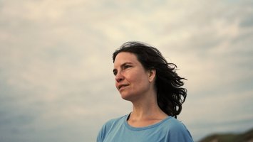 Autorin im Halbprofil, blaues Kurzarmshirt, wehende schulterlange, braune Haare, vor bedecktem Himmel