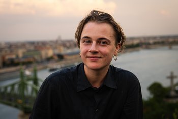 Autor*in vor See, kurze, leicht wellige braune Haare, goldener Ohrring links, schwarzes Hemd, lächelnd