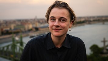 Autor*in vor See, kurze, leicht wellige braune Haare, goldener Ohrring links, schwarzes Hemd, lächelnd